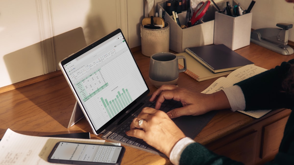 A person's hands typing on a laptop showing a spreadsheet with a cost breakdown chart, a coffee mug and notebook on the desk beside it