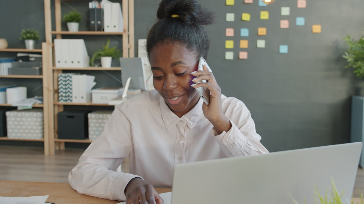 A professional talking on the phone at an office desk while looking at a laptop and a scheduling calendar
