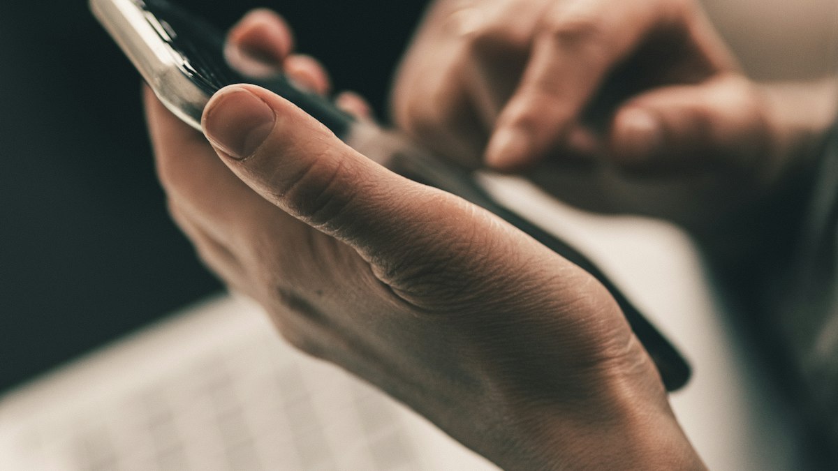 A person holding a smartphone above an open laptop keyboard, checking a website across devices