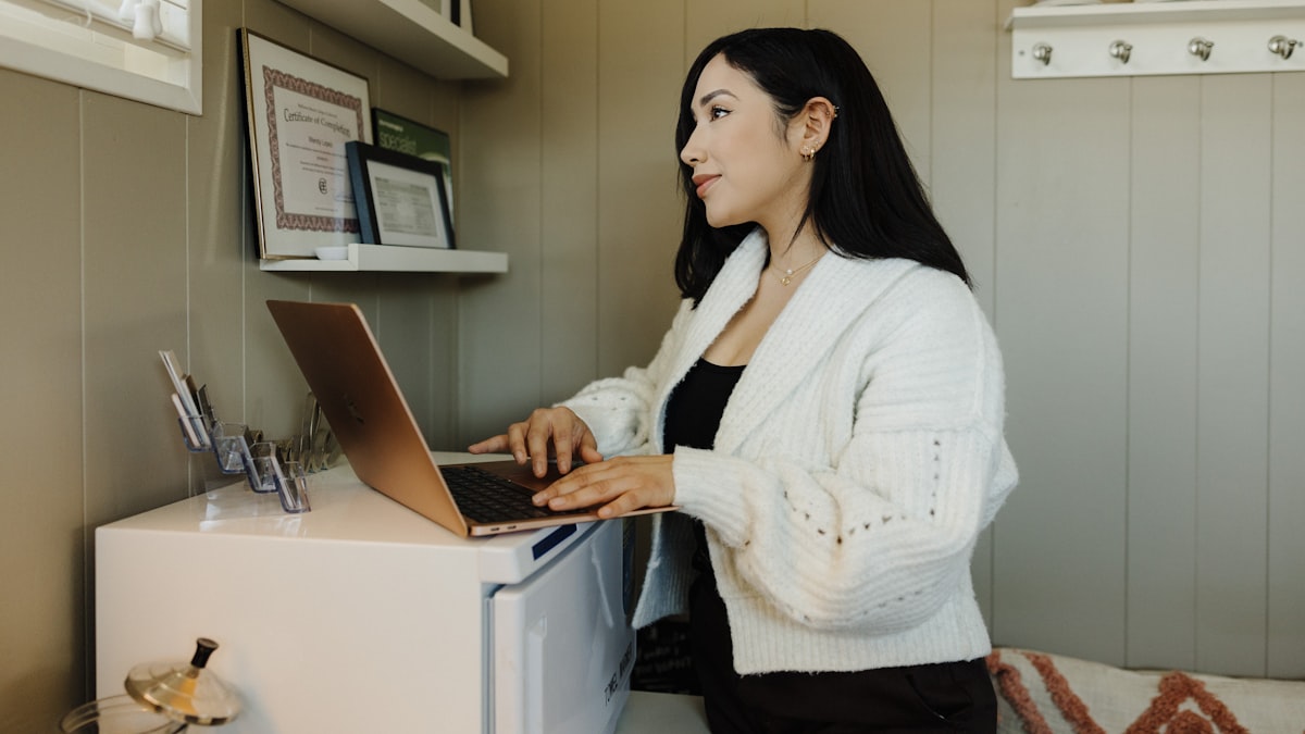 A small business owner standing at a counter, typing on a laptop next to a shelf holding a certificate and a framed photo, in a compact home-based shop
