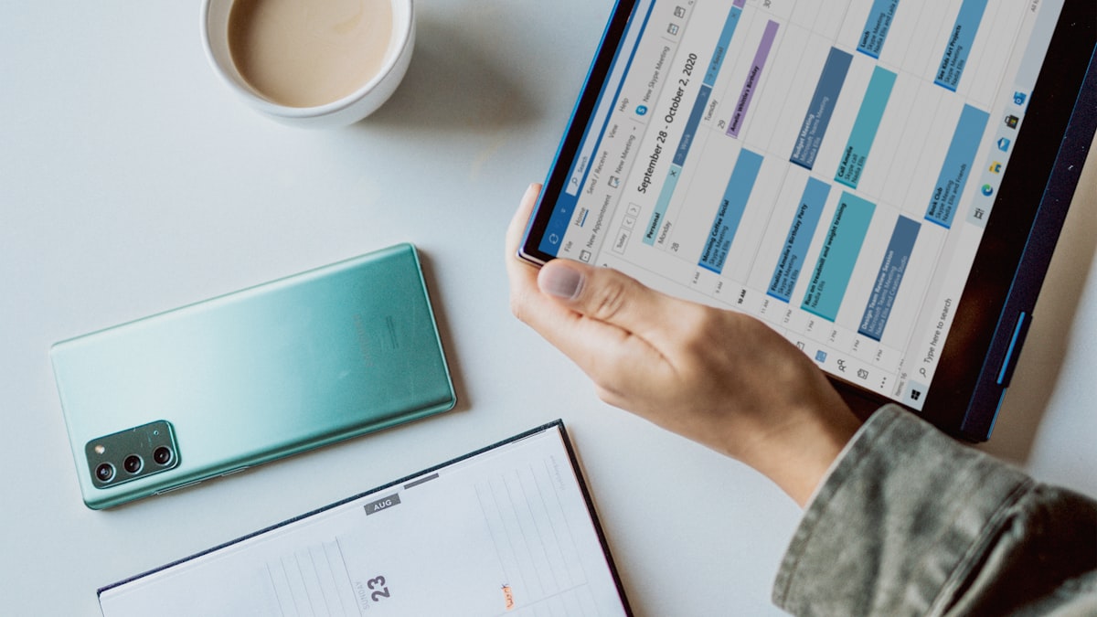 A person holding a tablet with a weekly schedule open, next to a phone and a paper planner
