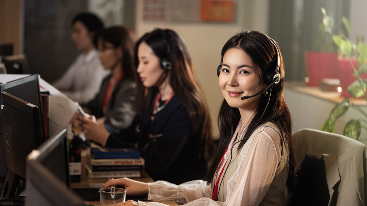 A customer service agent wearing a headset at a desktop computer in an office, with other agents working behind her